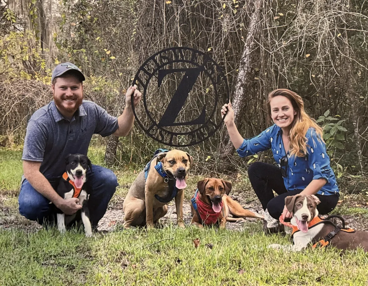 Megan and her husband kneeling in the woods with their four dogs, holding up a Zeisweiss family sign